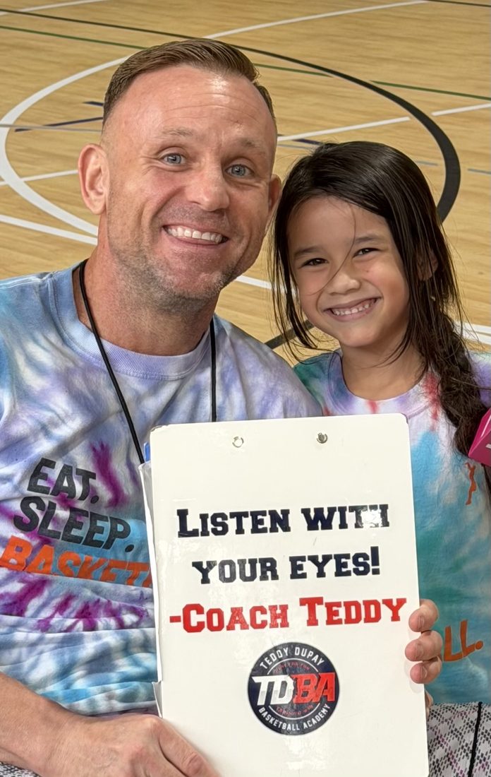 Coach Teddy at a TDBA camp with a young player, holding a clipboard that reads "Listen with your eyes! — Coach Teddy"